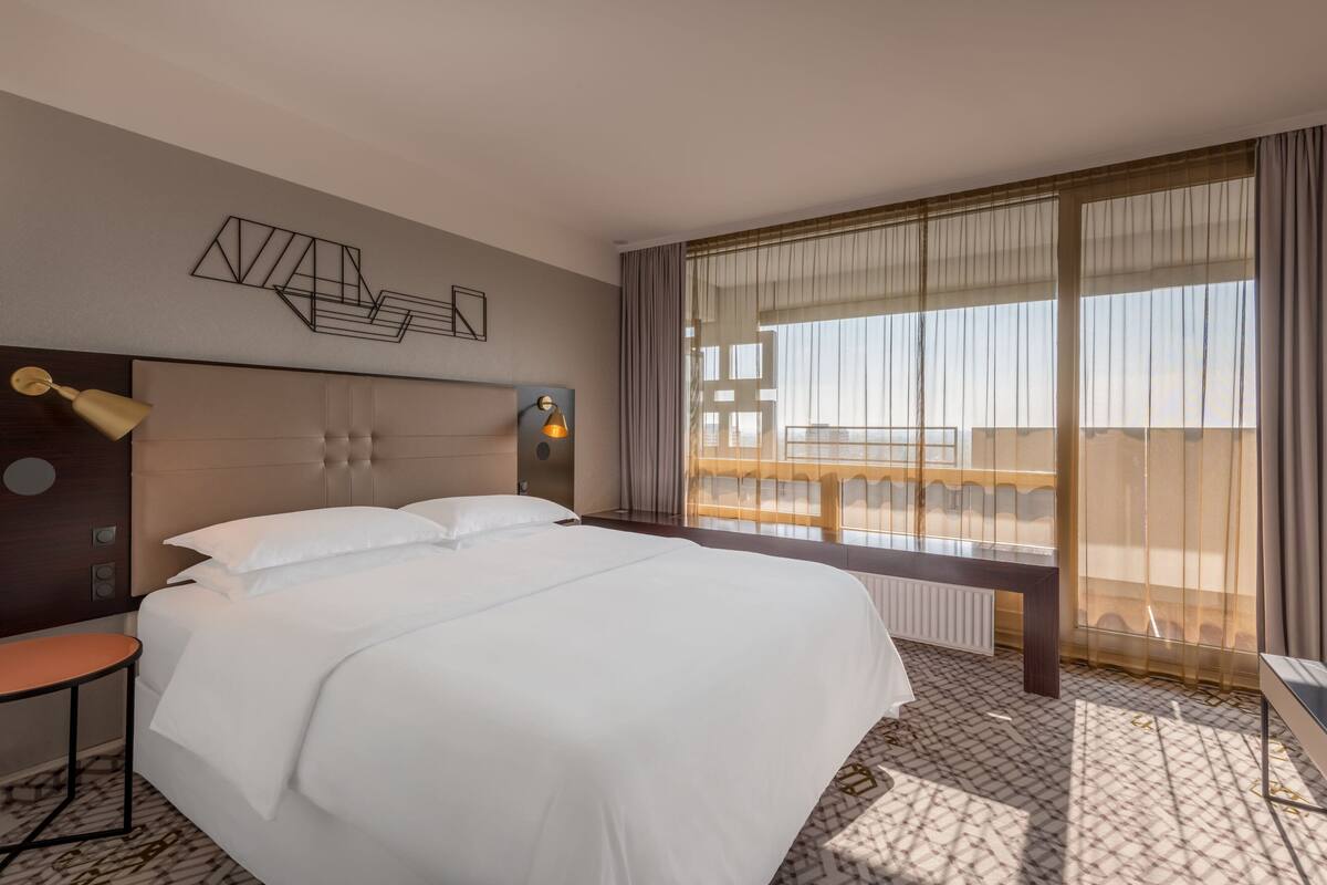 Ein Hotelzimmer mit einem großen Bett, einem Nachttisch, einer Lampe und Blick auf einen Balkon.
