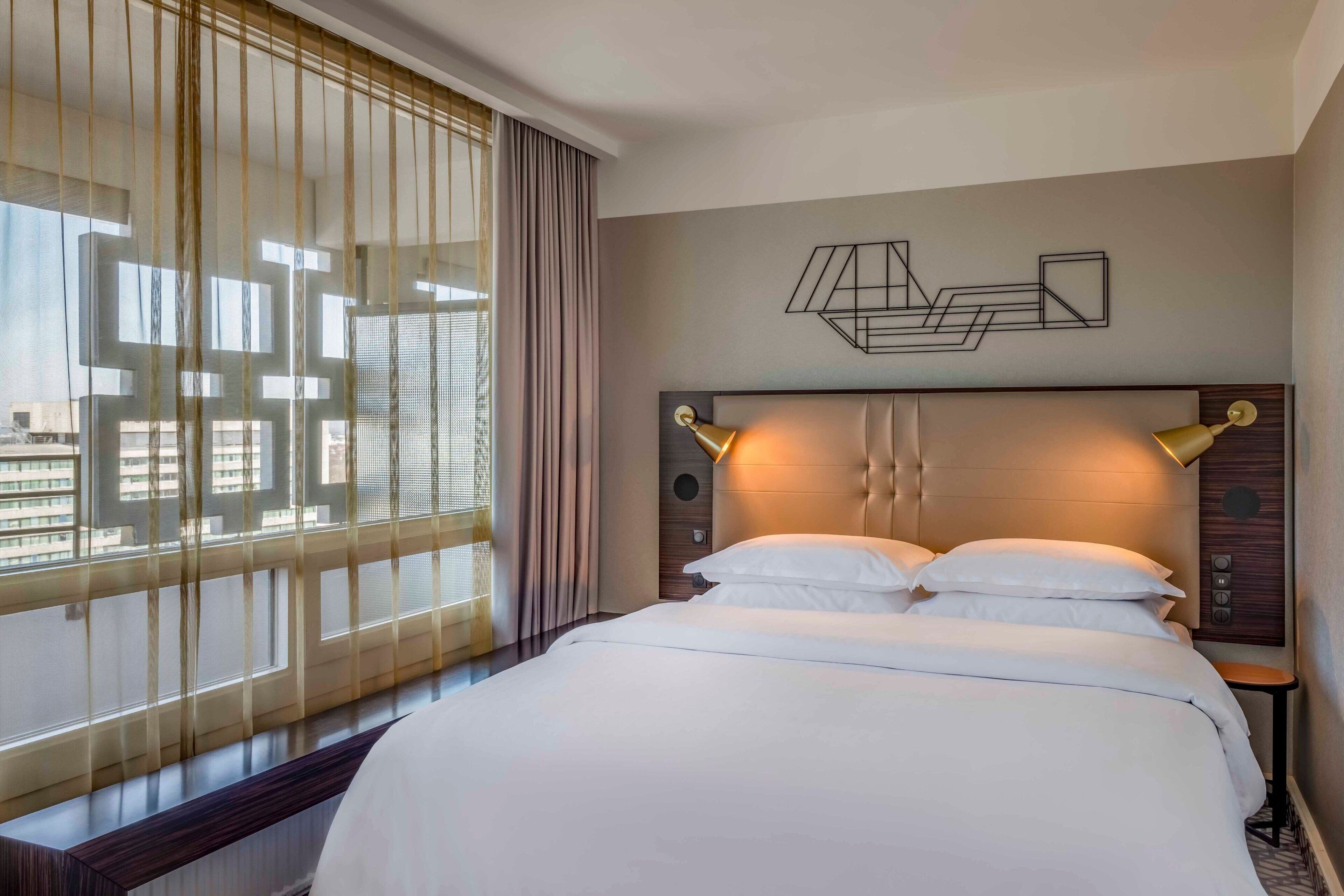 A hotel room with a large bed, a headboard with integrated lighting, a view of the city through sheer curtains, and a geometric wall art piece.