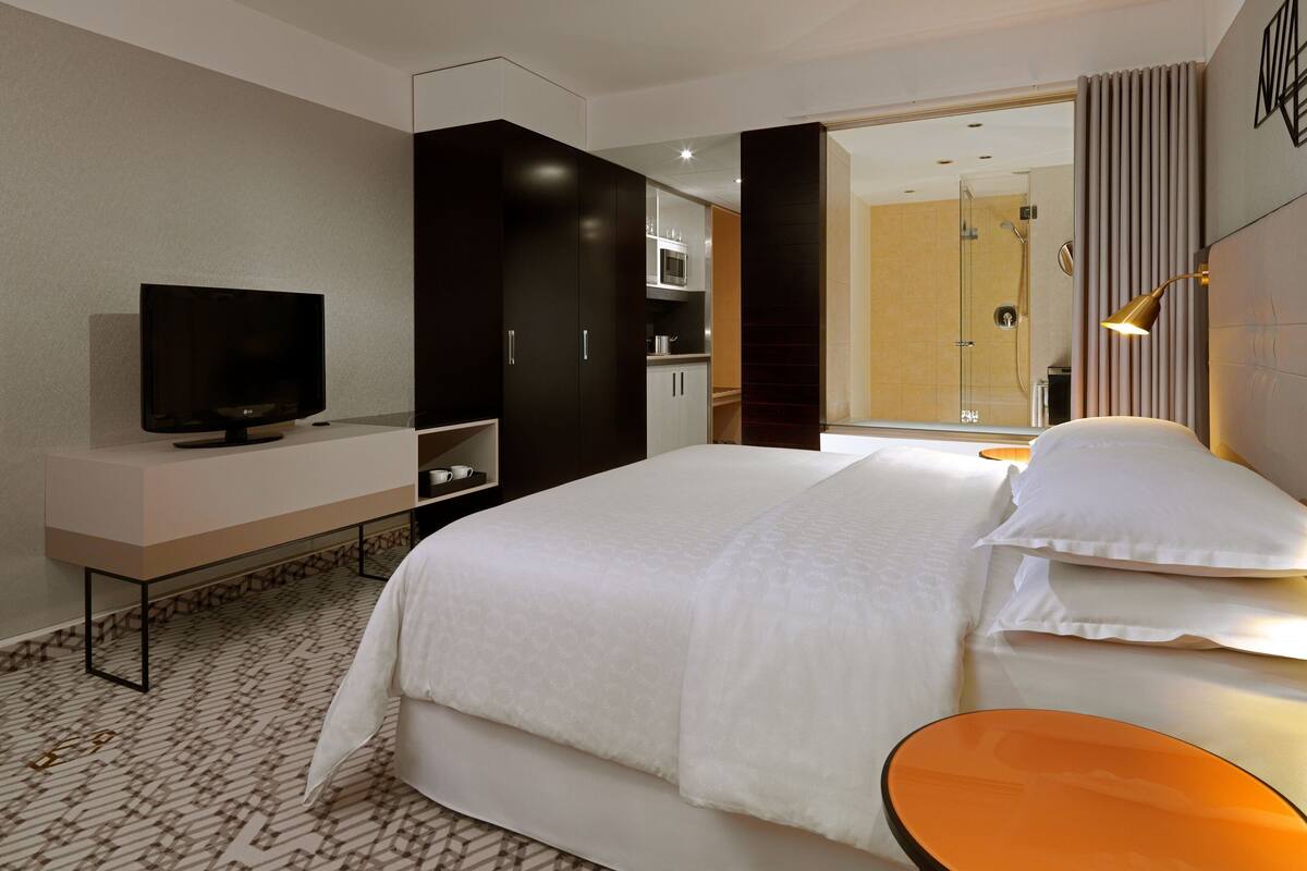 Ein modernes Hotelzimmer mit einem großen Bett, einem Fernseher, einer kleinen Küchenzeile und einem durchgegangenen Tür erkennbaren Badezimmer.