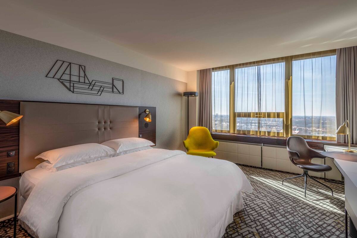 Ein Hotelzimmer mit einem großen Bett, einem Schreibtisch, einem Sessel und Blick auf die Stadt durch große Fenster.