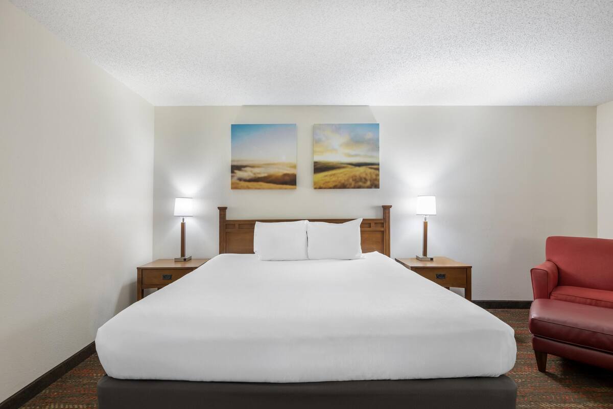 Un cuarto de hotel con una cama grande, dos mesitas de noche con lámparas, una silla roja y dos cuadros de paisajes en la pared.