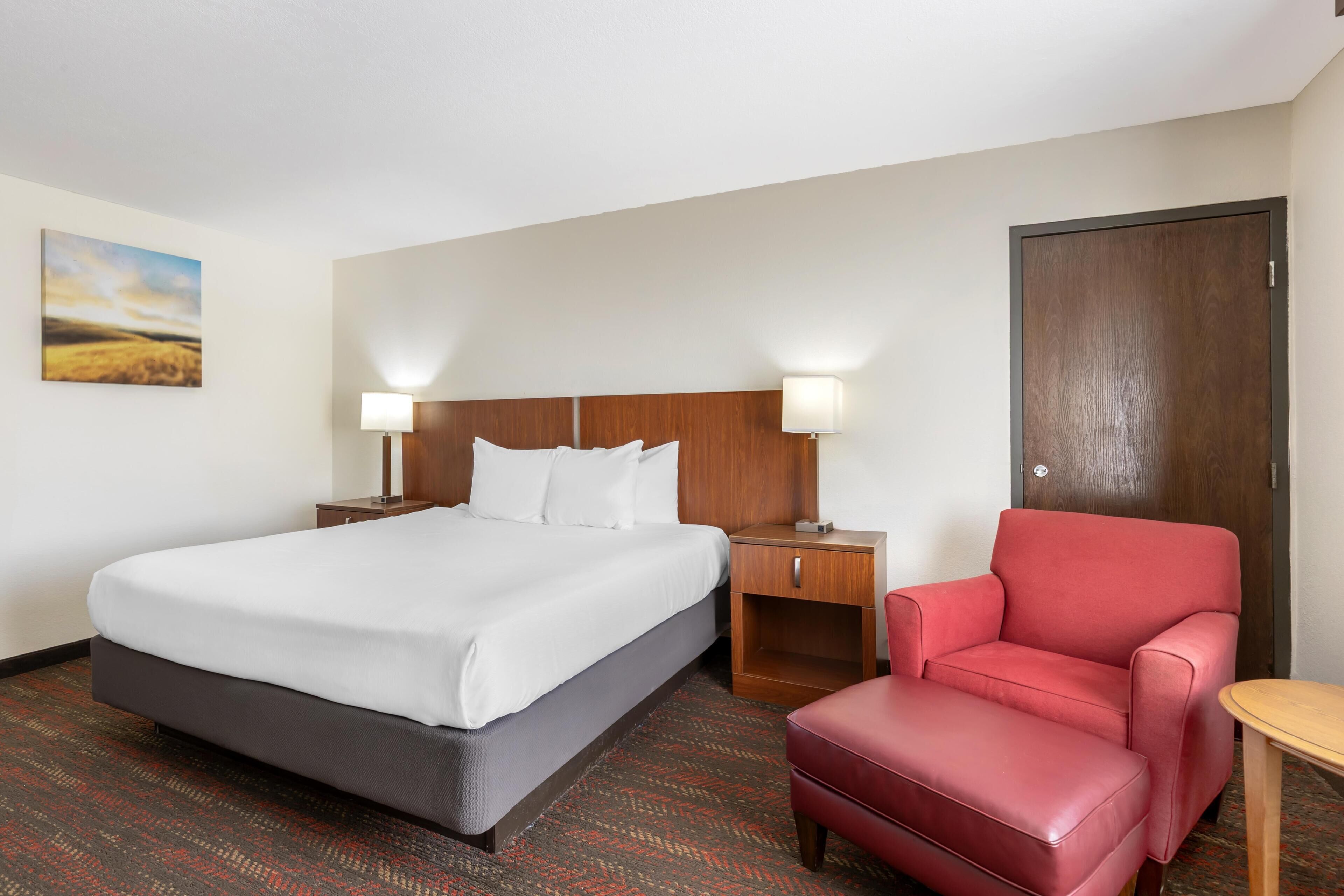 A hotel room with a bed, two bedside tables with lamps, a red chair, a wooden door, and a painting on the wall.