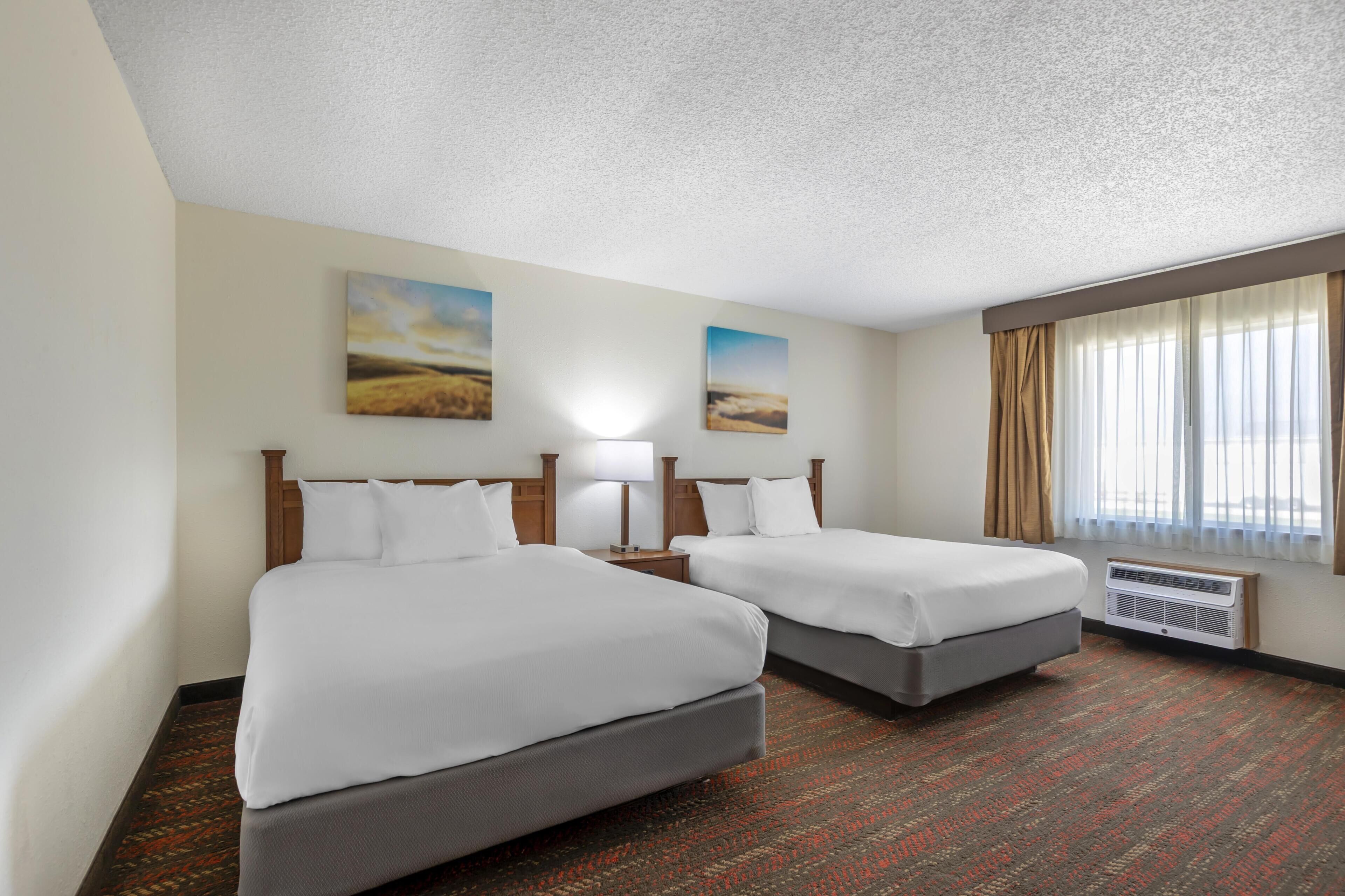 Habitación de hotel con dos camas, una ventana con cortinas, aire acondicionado de pared y dos cuadros.