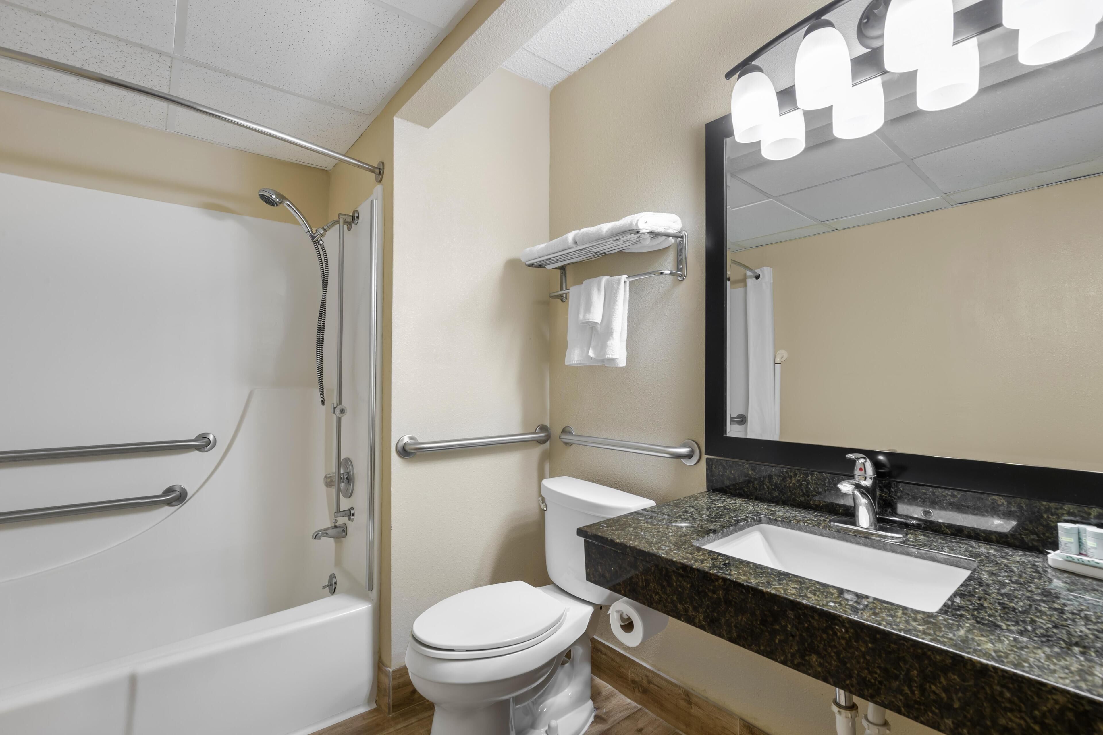 Une salle de bain équipée d’une baignoire, de toilettes et d’un lavabo, ainsi que de barres d’appui et d’un miroir avec éclairage intégré.
