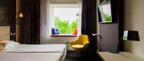 Habitación de hotel con cama, escritorio, silla, televisor y ventana con vista a follaje.