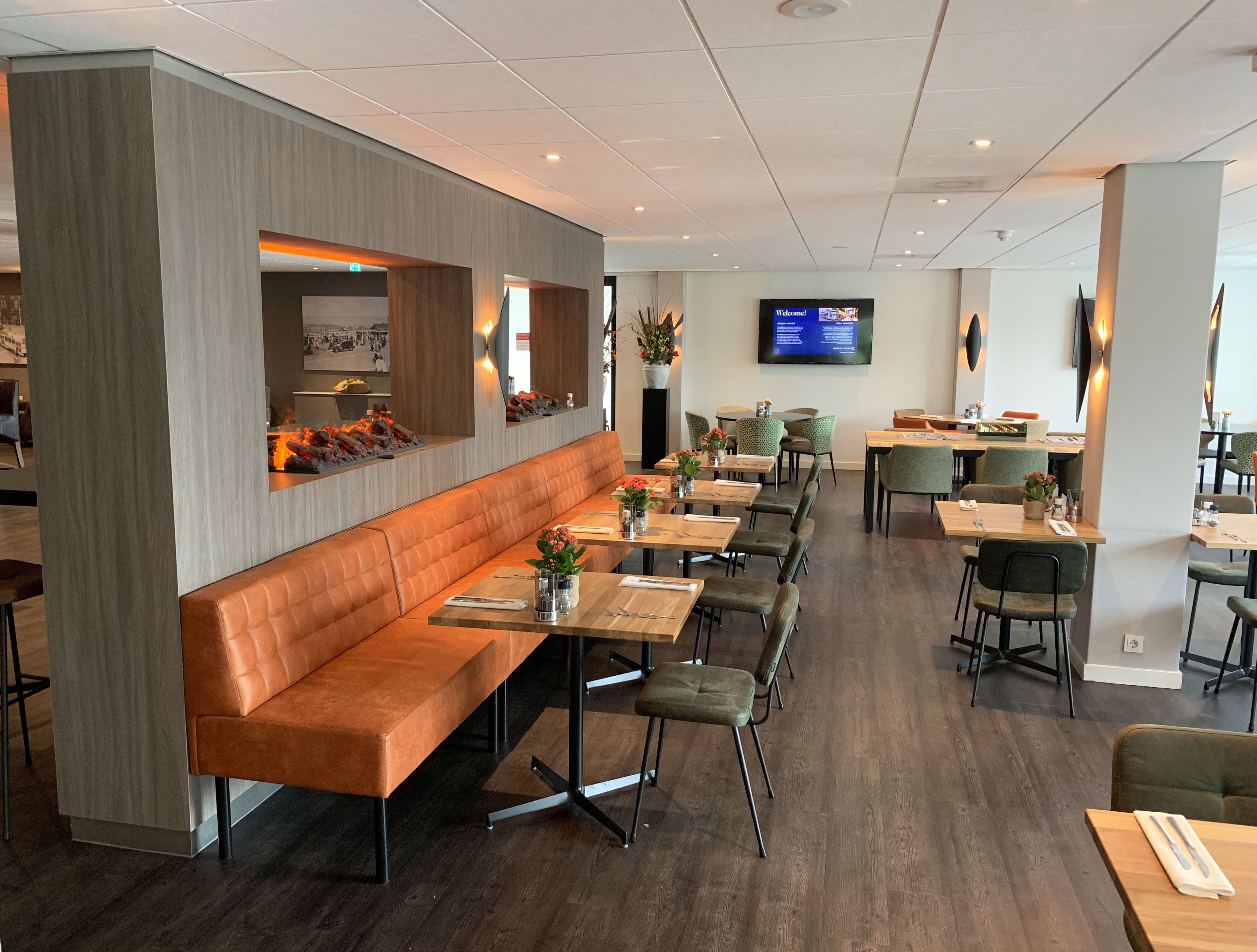 A modern restaurant with a long wooden bench seating area, multiple tables with chairs, and a flat-screen TV mounted on the wall.