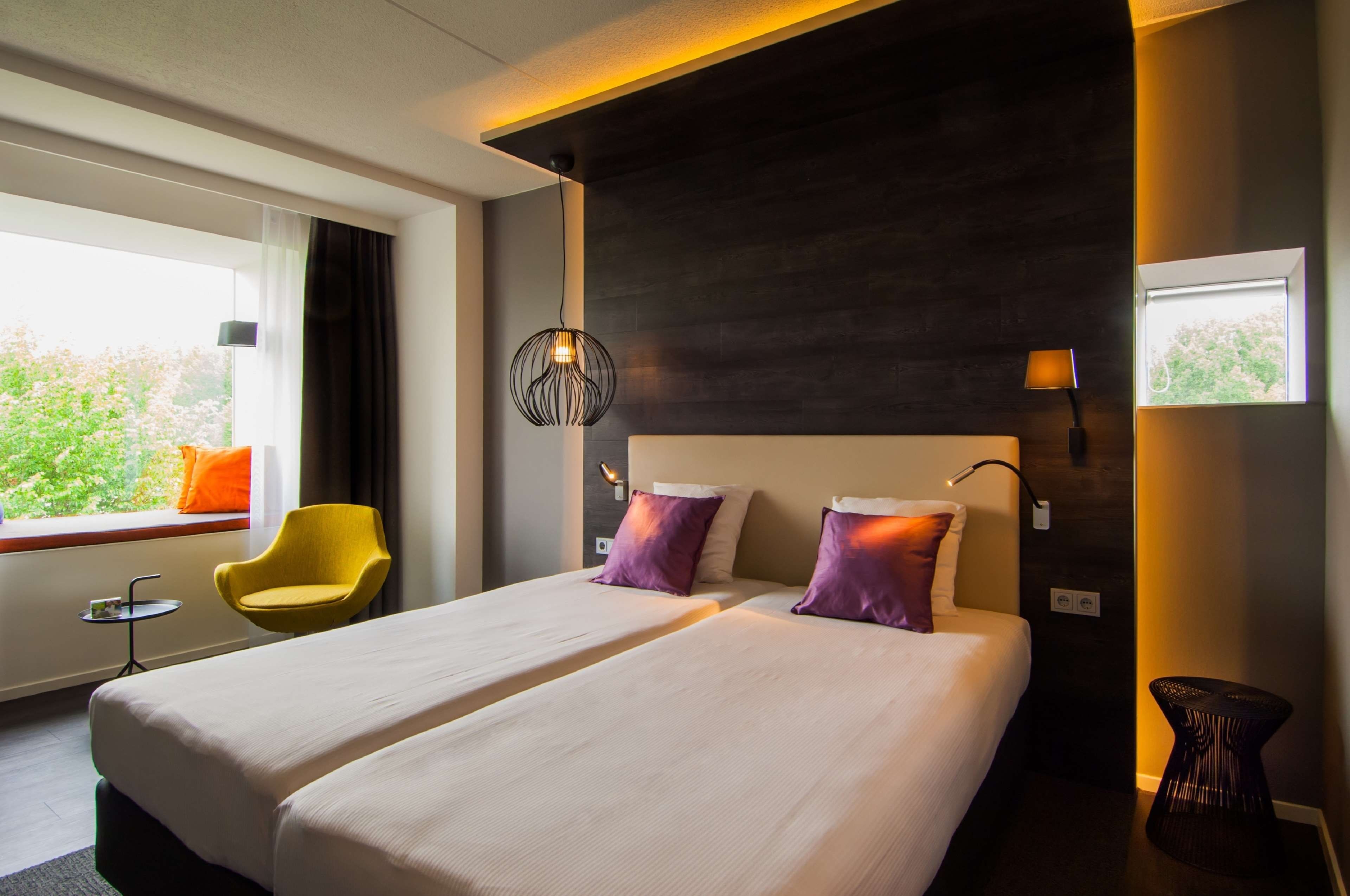 Une chambre d’hôtel avec un grand lit, une chaise, une petite table et une vue sur la verdure environnante.