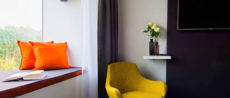 Una habitación de hotel moderna con un ventanal grande, un sillón amarillo, un televisor de pantalla plana y una mesita de noche con un libro y un jarrón de flores.