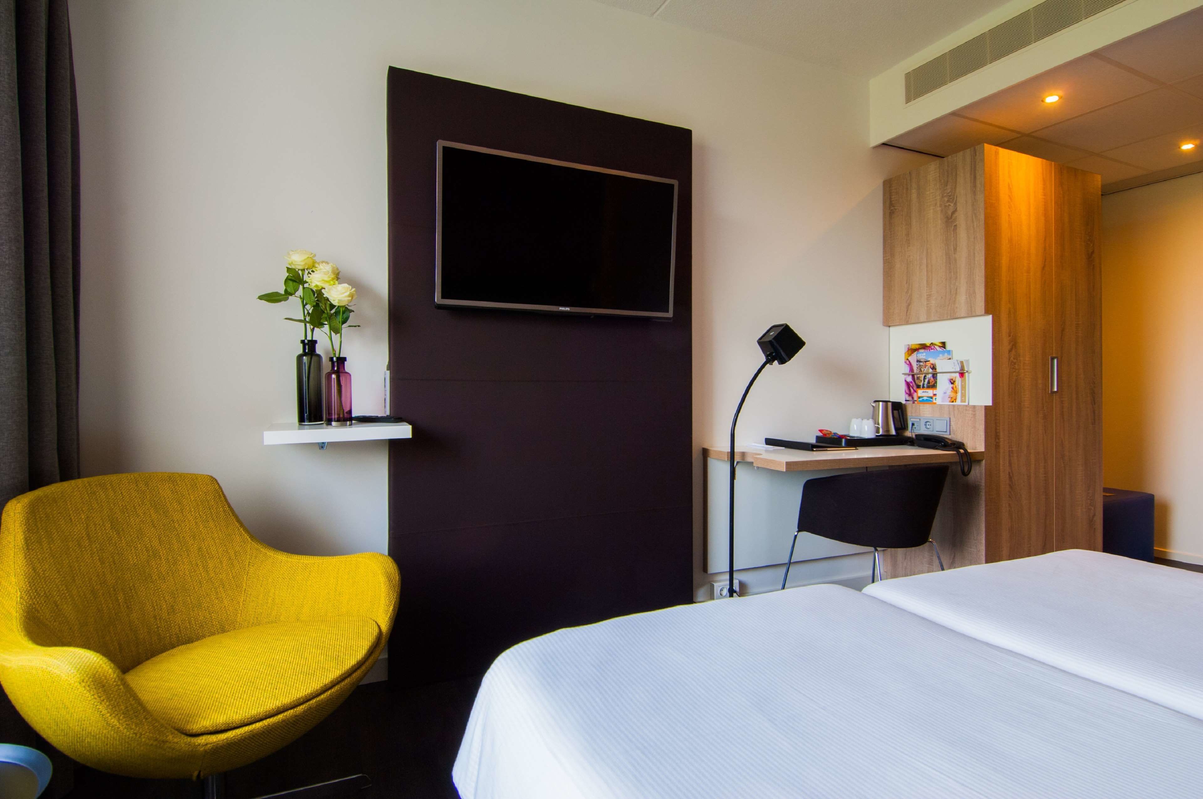 A hotel room with a bed, a yellow armchair, a desk with a computer, a flat-screen TV, and a small shelf with flowers.