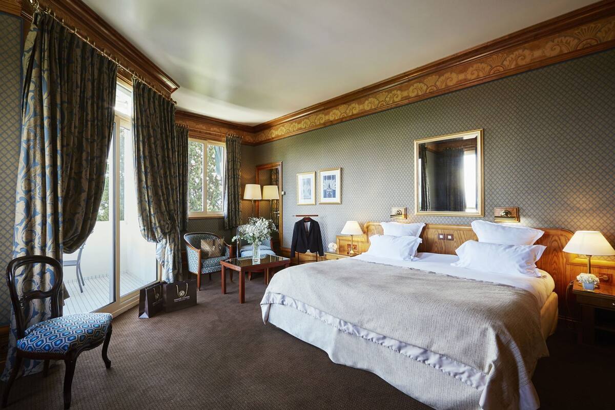 A hotel room with a large bed, a sitting area with a chair and table, a wardrobe, and a window with curtains.