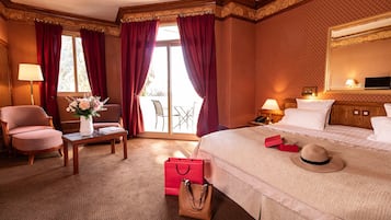 Una habitación de hotel con una cama grande, una silla, una mesa con un florero y una bolsa de compras roja, además de un sombrero sobre la cama.