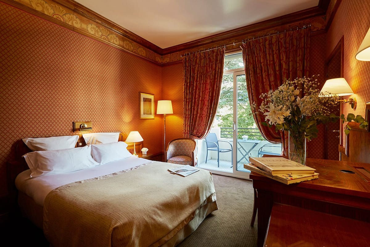 A hotel room with a large bed, two bedside tables with lamps, a chair, a desk with a lamp, and a view of the outdoors through a sliding door.