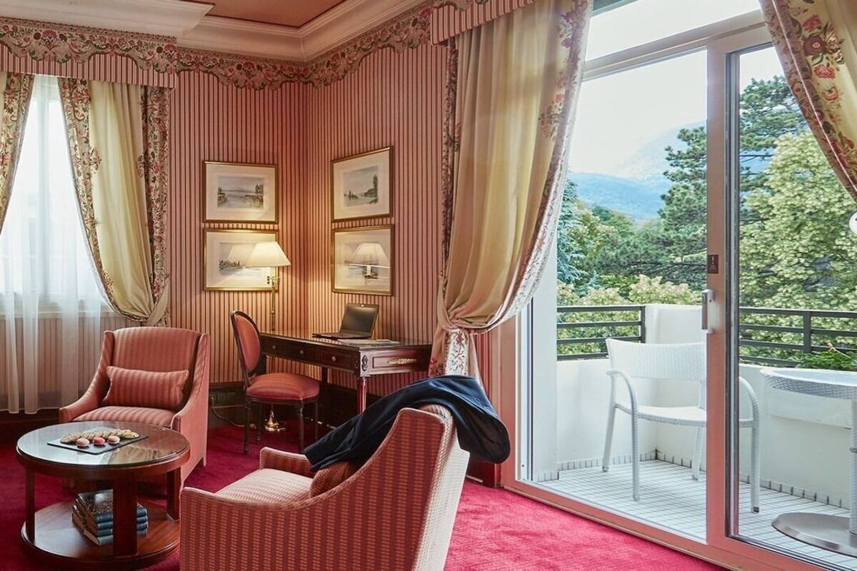 A room with striped wallpaper, a red sofa, a round coffee table, a desk with a lamp, and a balcony with white chairs.