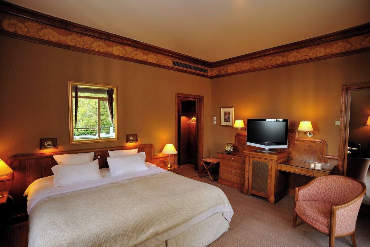 A hotel room with a large bed, a desk with a television, a chair, and a window with a view of greenery.