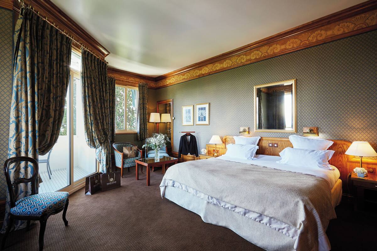 A hotel room with a large bed, a sitting area with a chair and table, a dresser, and a window with curtains.