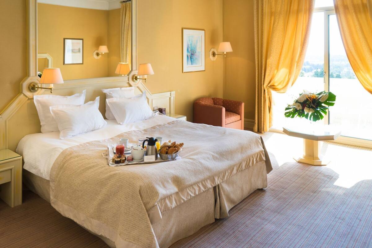A hotel room with a large bed, bedside tables, a chair, a small table with a vase, and a window with curtains.