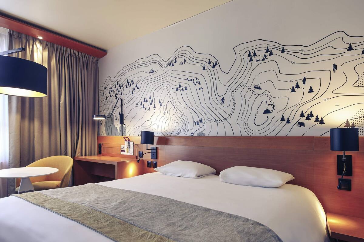 A hotel room with a large bed, a desk, two chairs, and a wall with a topographic map design.