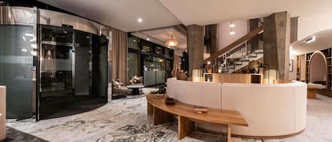 Un interior moderno con piso de mármol, un sofá curvo, un banco de madera y una escalera con barandilla de madera.