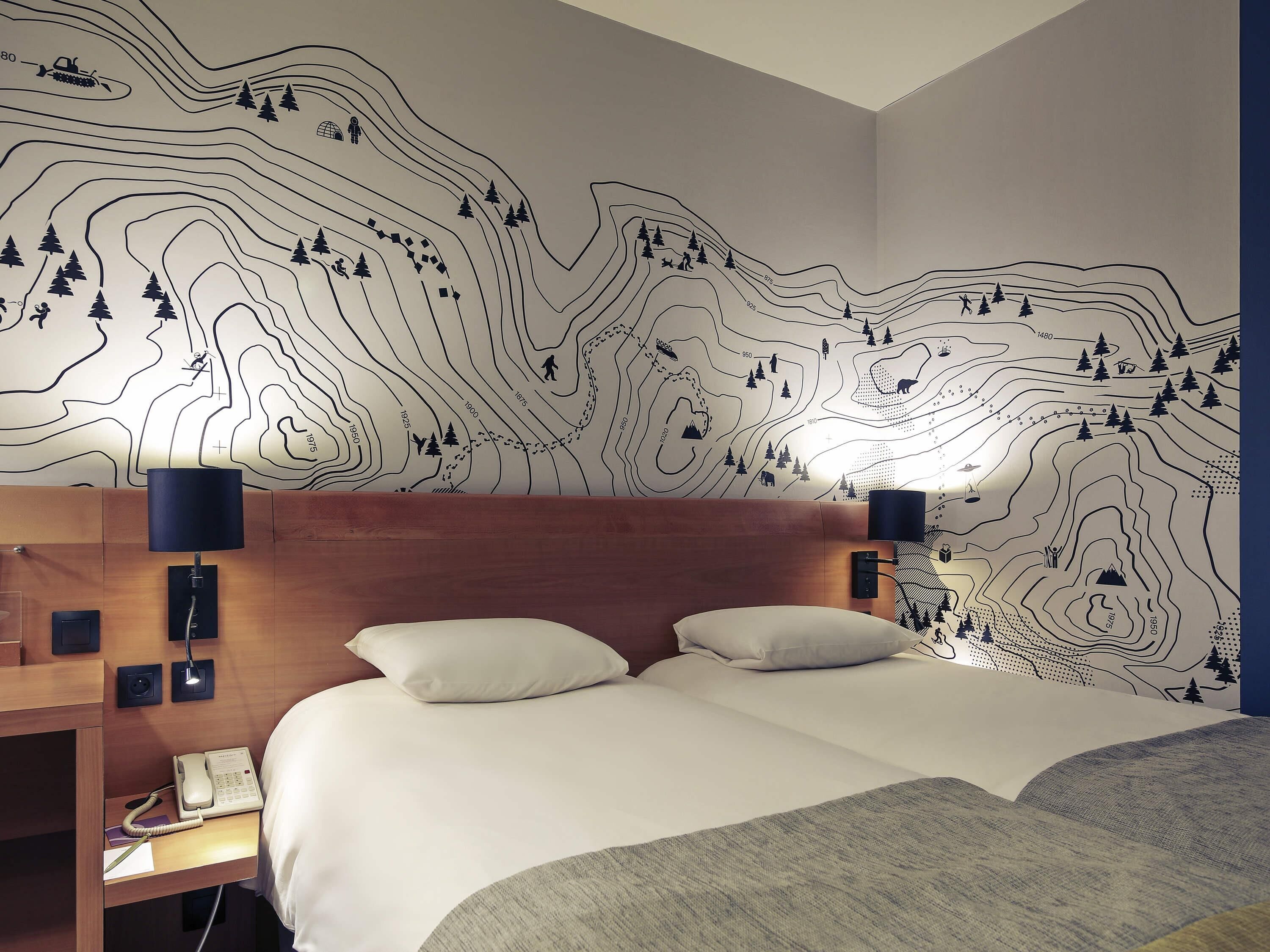 Une chambre d’hôtel avec une grande fresque murale représentant un paysage de montagne, une tête de lit en bois, deux oreillers blancs, une table de chevet avec une lampe et un téléphone.