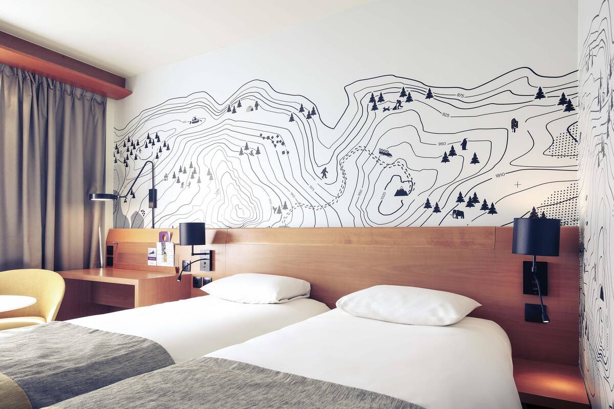 A hotel room with two beds, a wooden headboard, a desk, a chair, and a wall with a topographic map design.