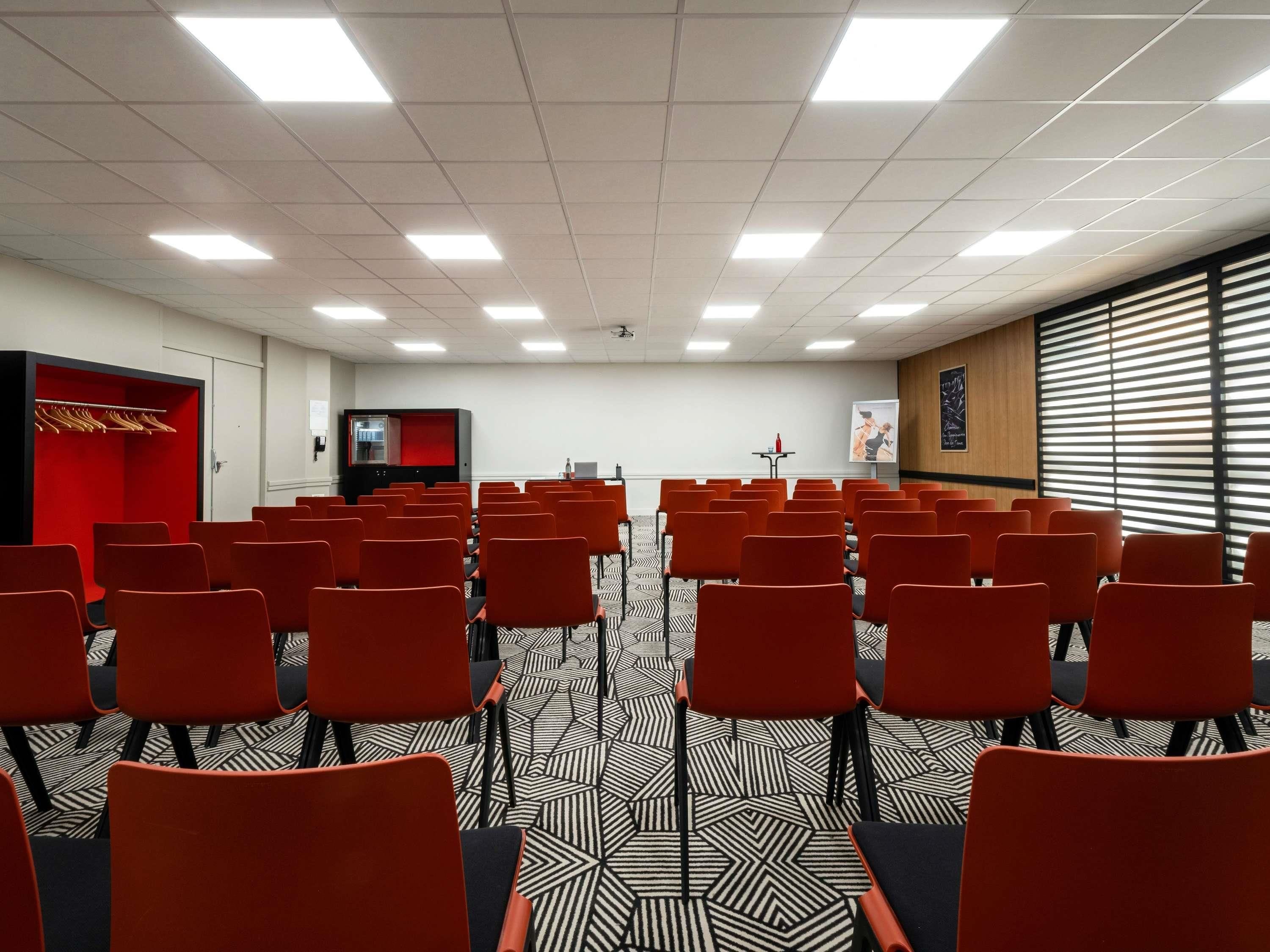 Une salle de conférence avec des rangées de chaises, un écran de projection et un espace détente avec des sièges rouges.