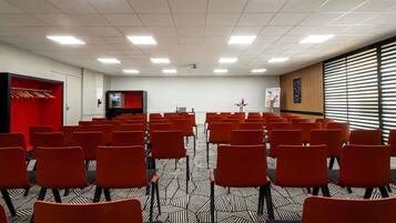 Une salle de conférence avec des rangées de chaises, un écran de projection et un espace détente avec des sièges rouges.