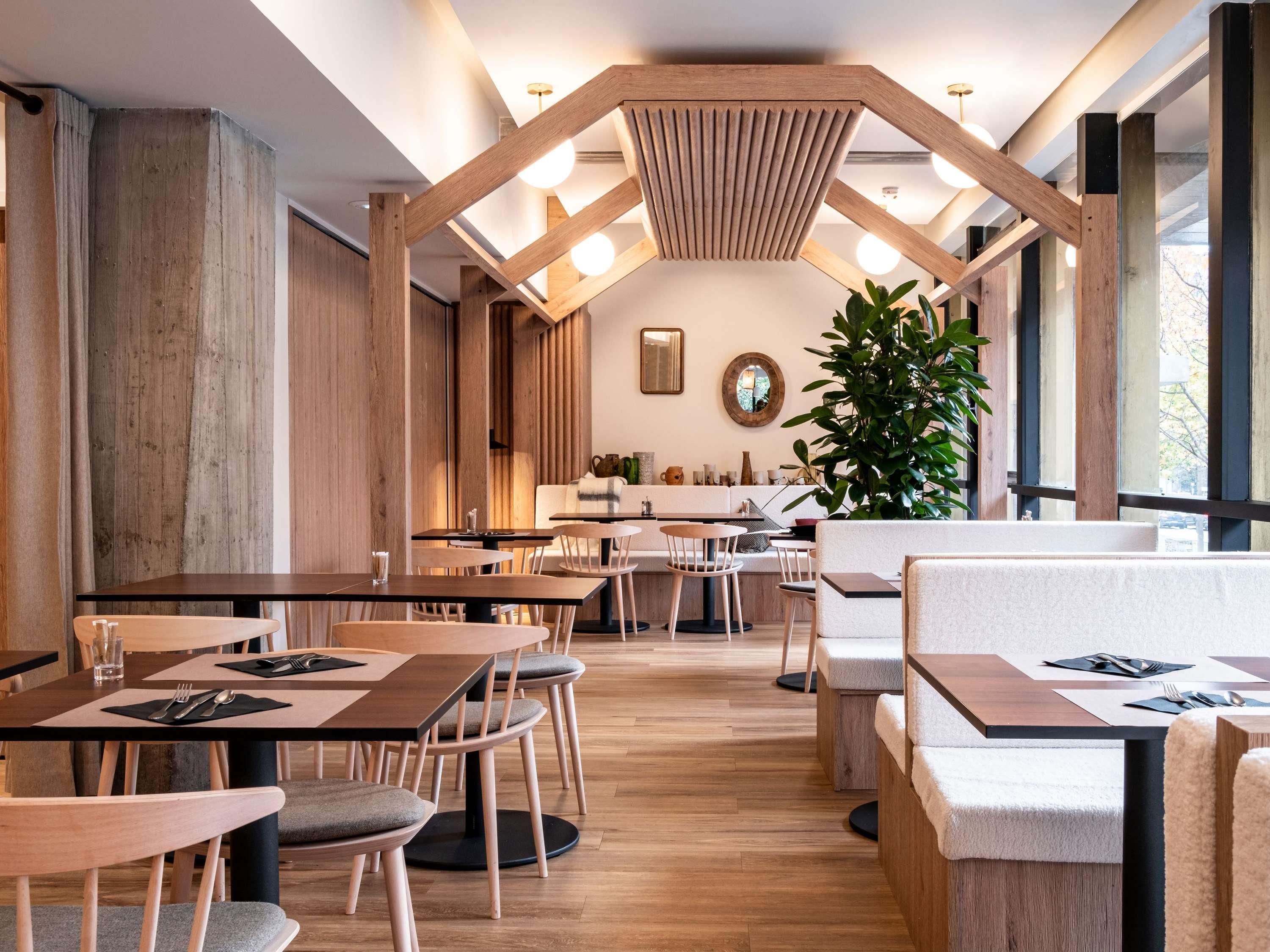 Un interior de restaurante moderno con vigas de madera, mesas redondas y una planta grande en maceta.