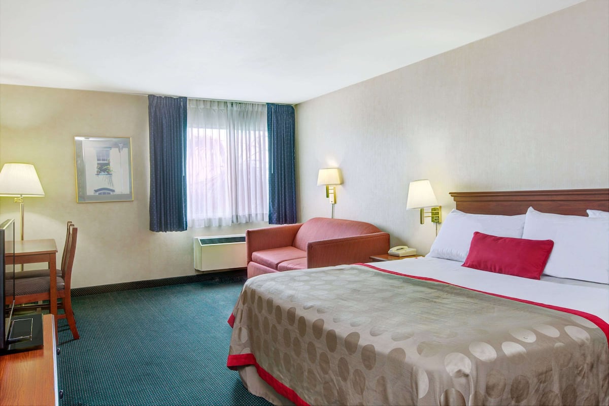 A hotel room with a bed, a desk, a chair, two lamps, a window with curtains, and a framed picture on the wall.