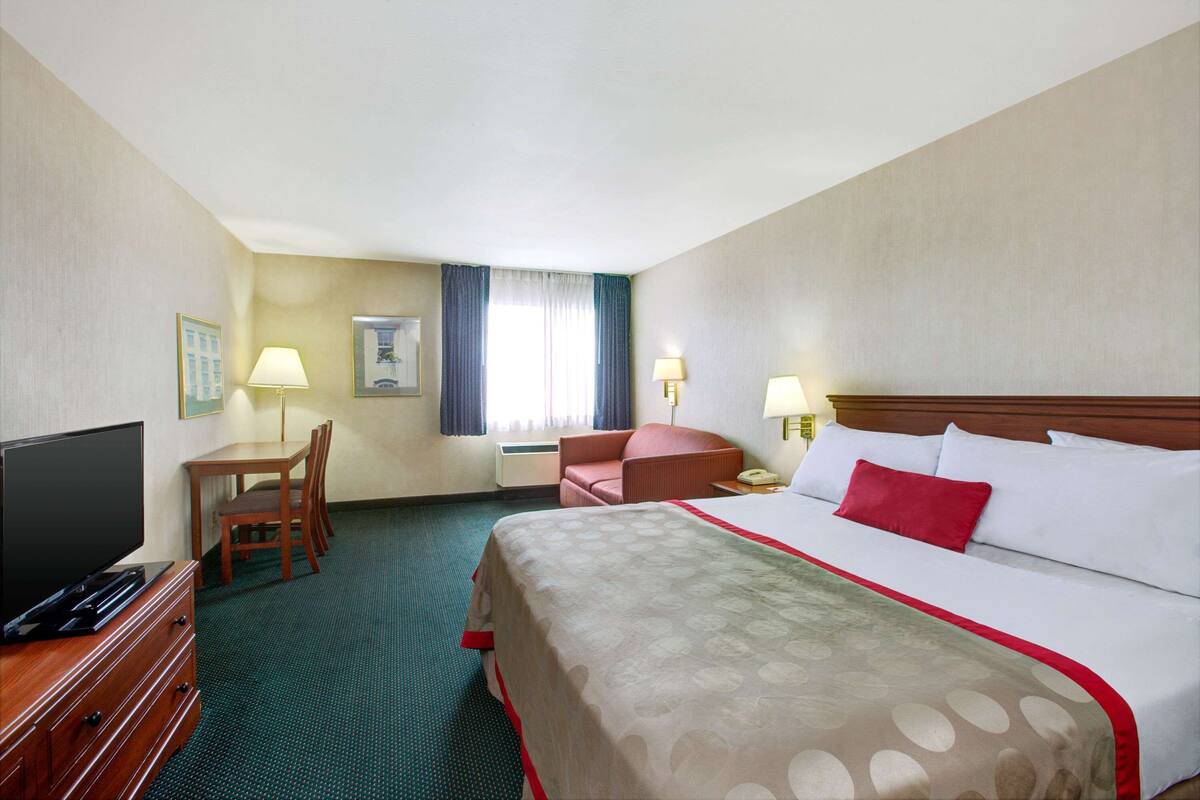 A hotel room with a bed, a desk with a chair, a television, and a window with curtains.