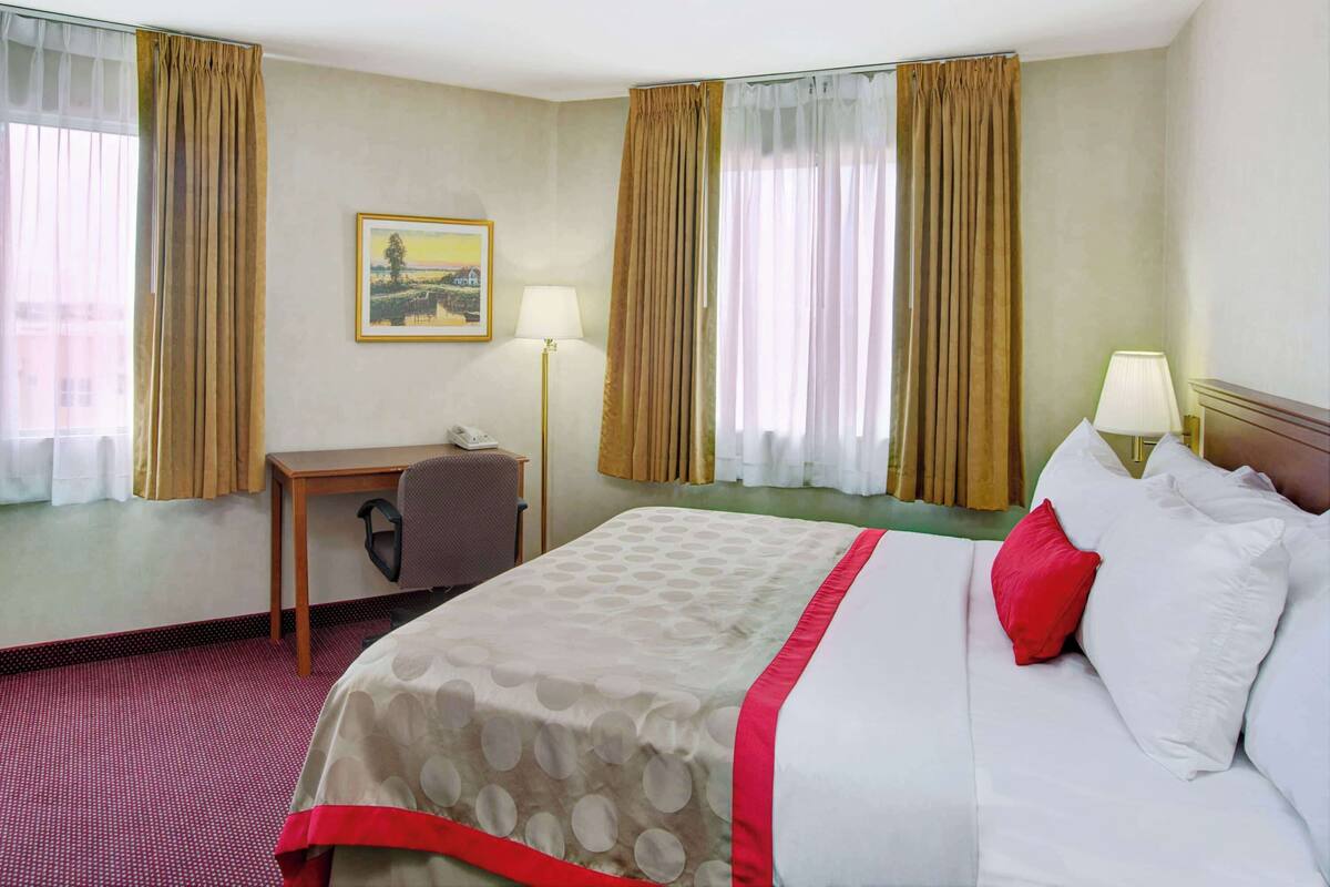 A hotel room with a bed, a desk, two lamps, and a painting on the wall.