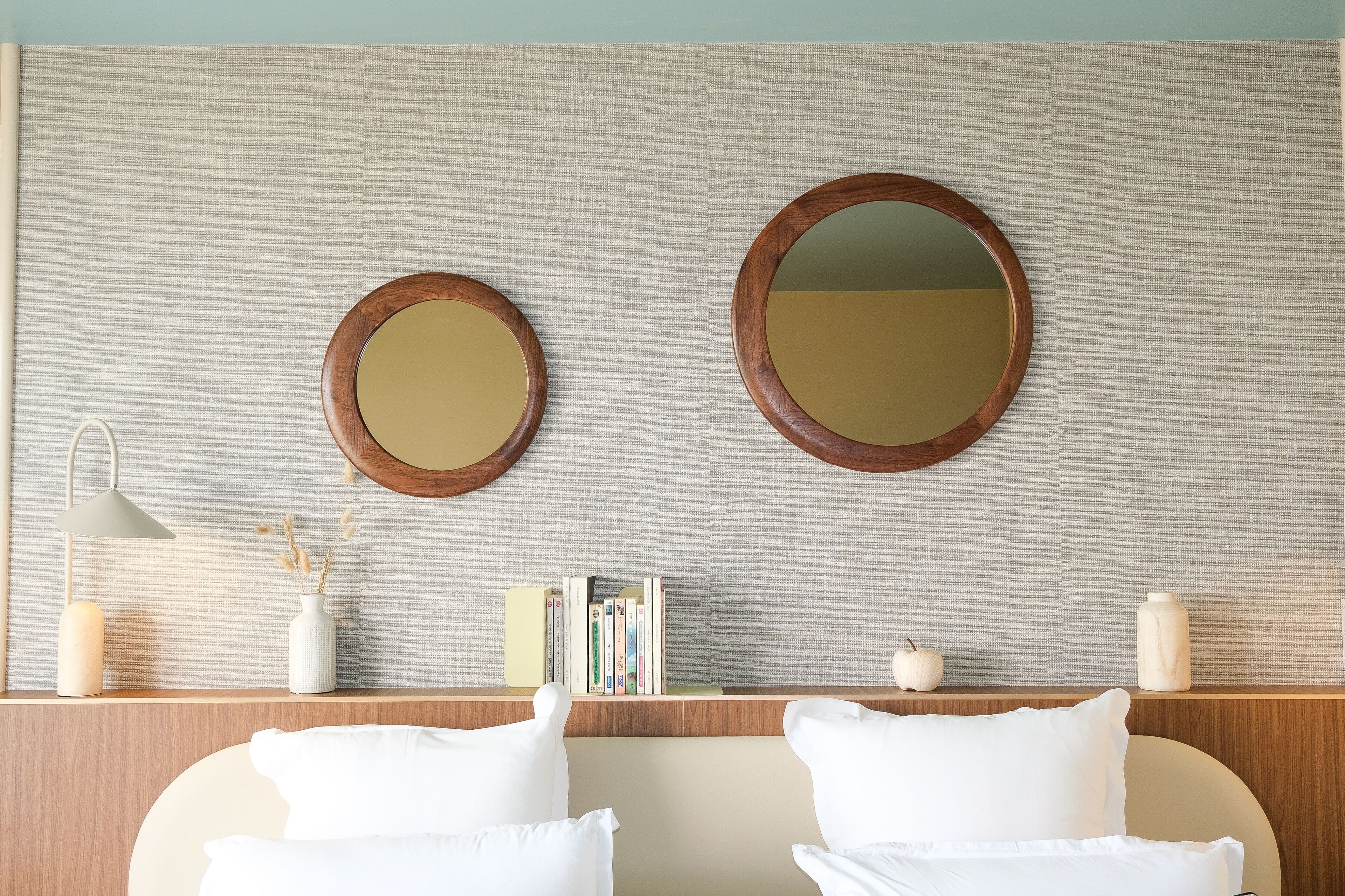 A neatly arranged hotel room with two round mirrors, a bedside lamp, a small bookshelf, and a wooden headboard.
