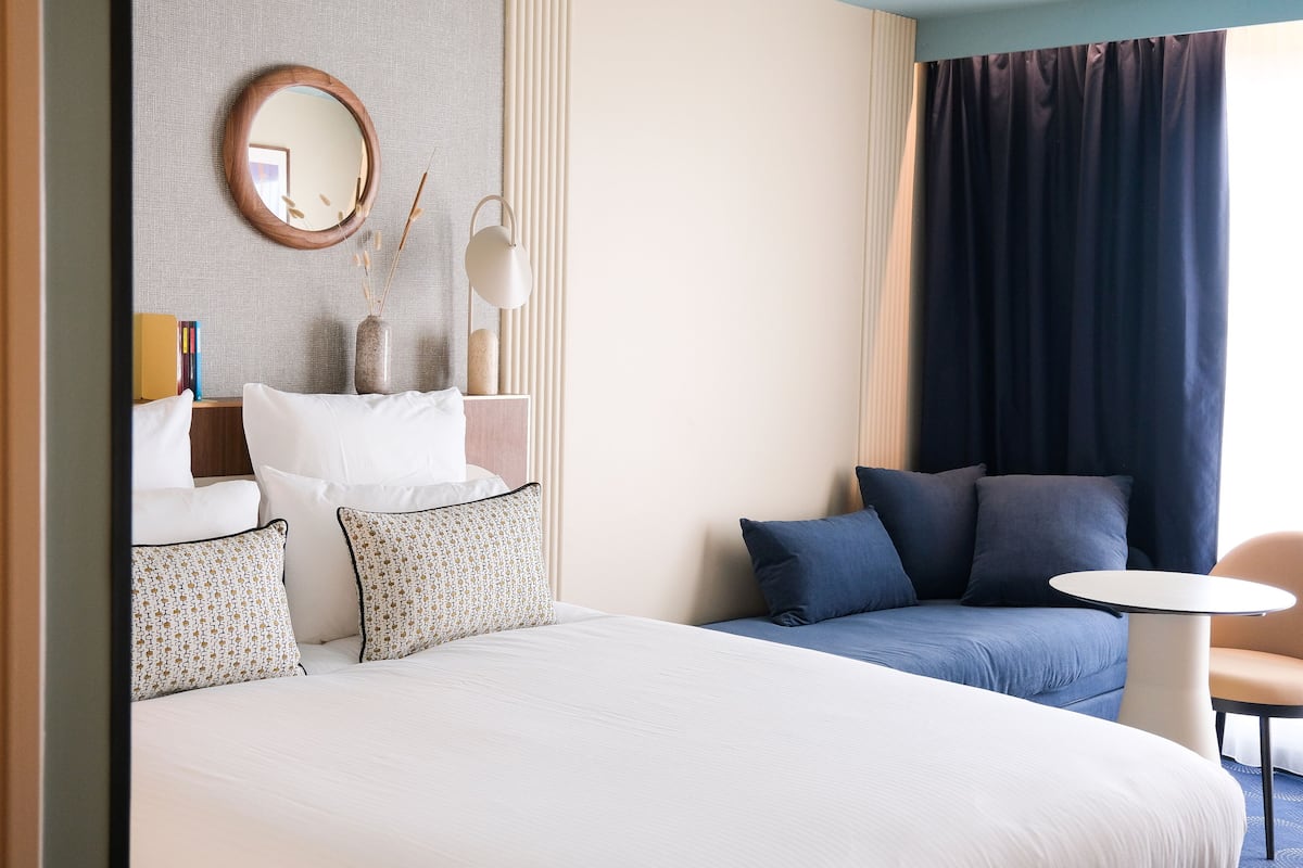 Habitación de hotel con una cama grande, un sofá, una mesita y una silla. Hay un espejo en la pared y un florero con plantas.