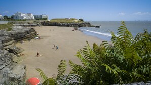Une plage bordée de falaises rocheuses, avec une étendue de sable et quelques bâtiments visibles au loin.