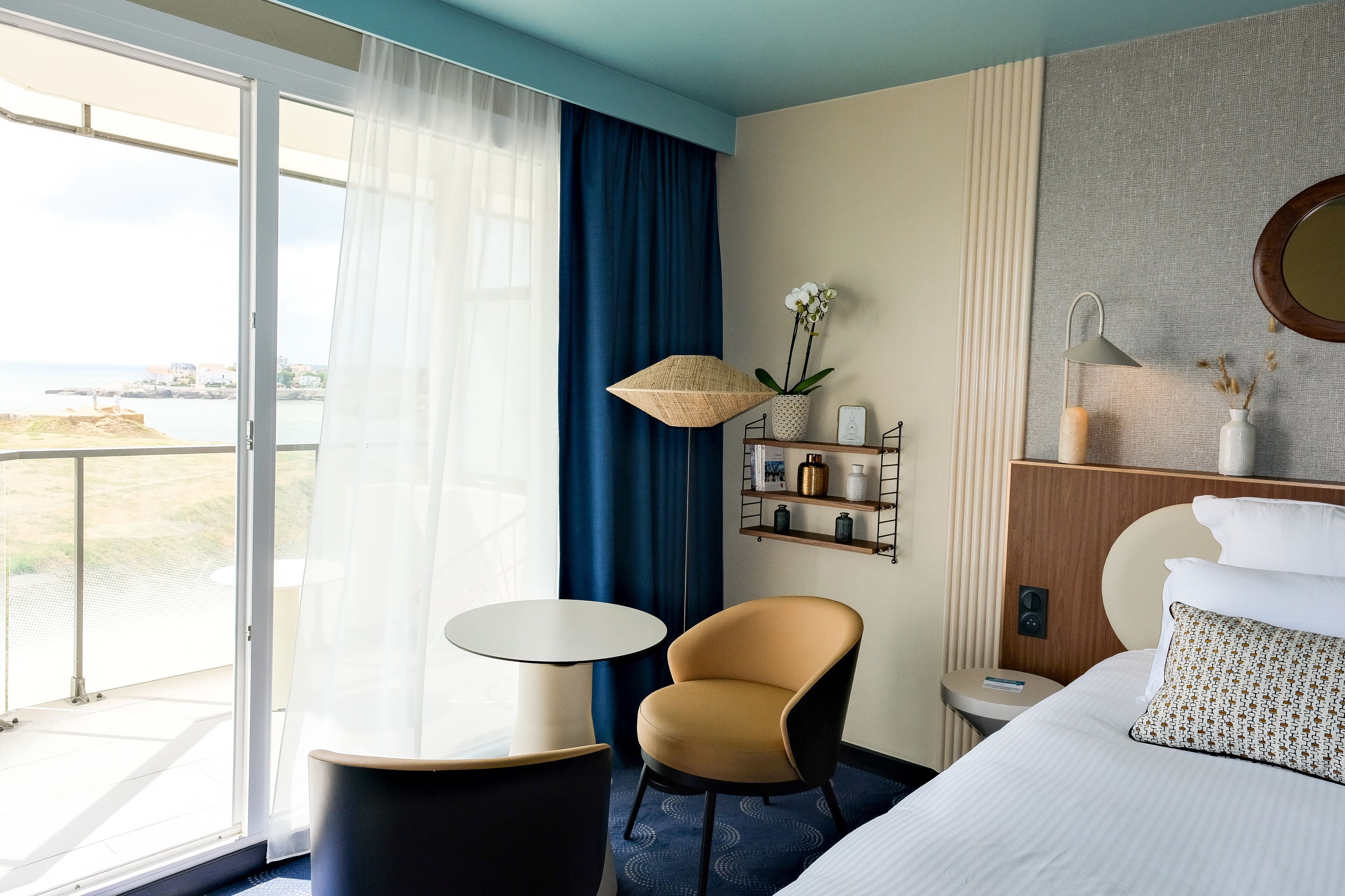 A hotel room with a bed, a chair, a small table, a nightstand, a lamp, a shelf with decorative items, and a view of the sea through large windows.
