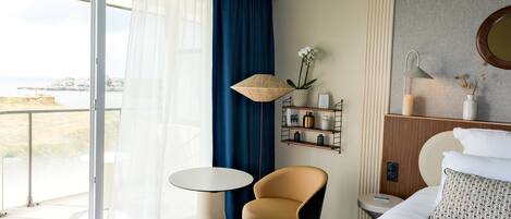 A hotel room with a bed, a chair, a small table, a nightstand, a lamp, a shelf with decorative items, and a view of the sea through large windows.