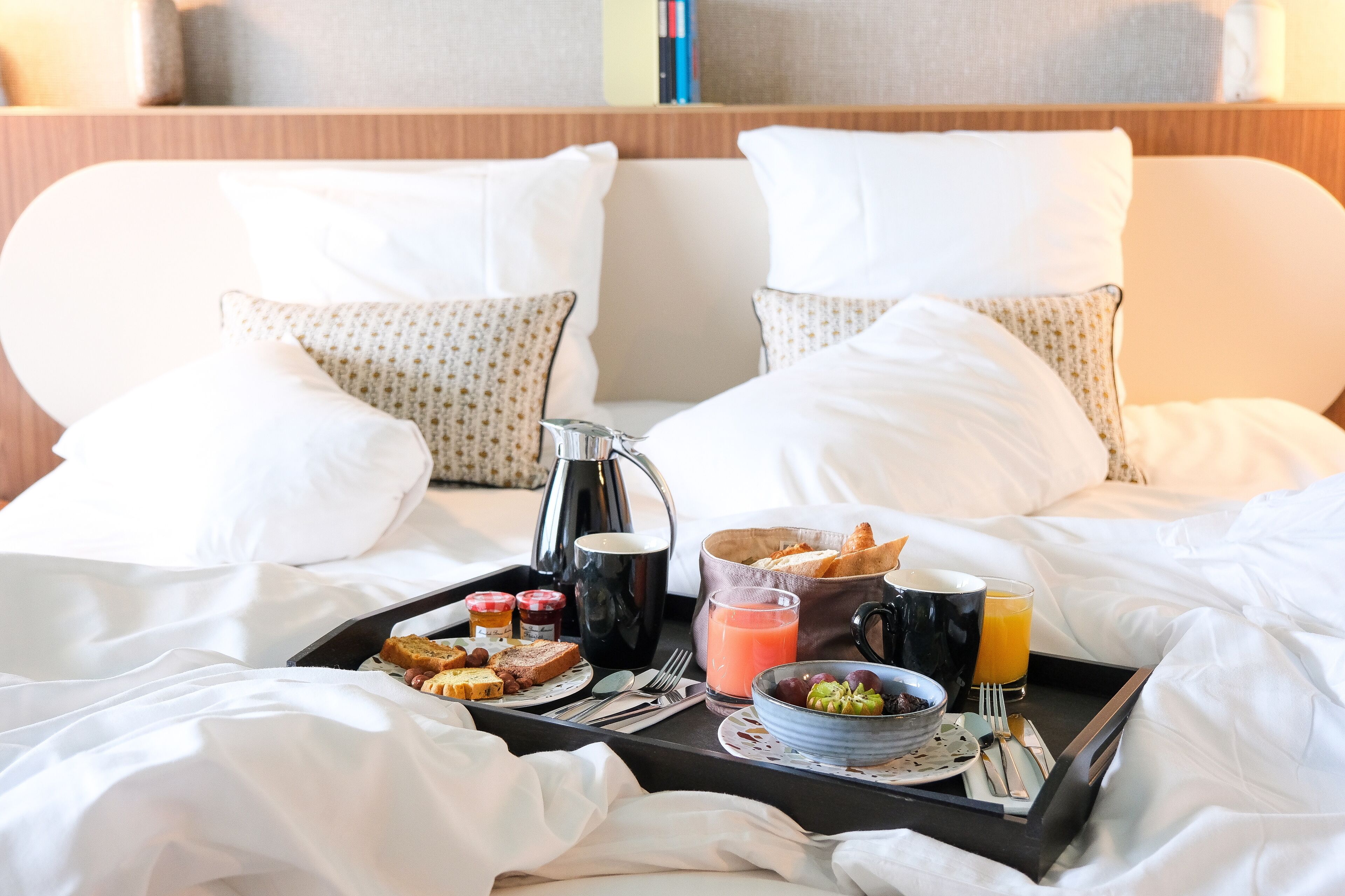 A neatly made bed with white linens, a tray with breakfast items including toast, jam, a coffee pot, and a glass of orange juice.