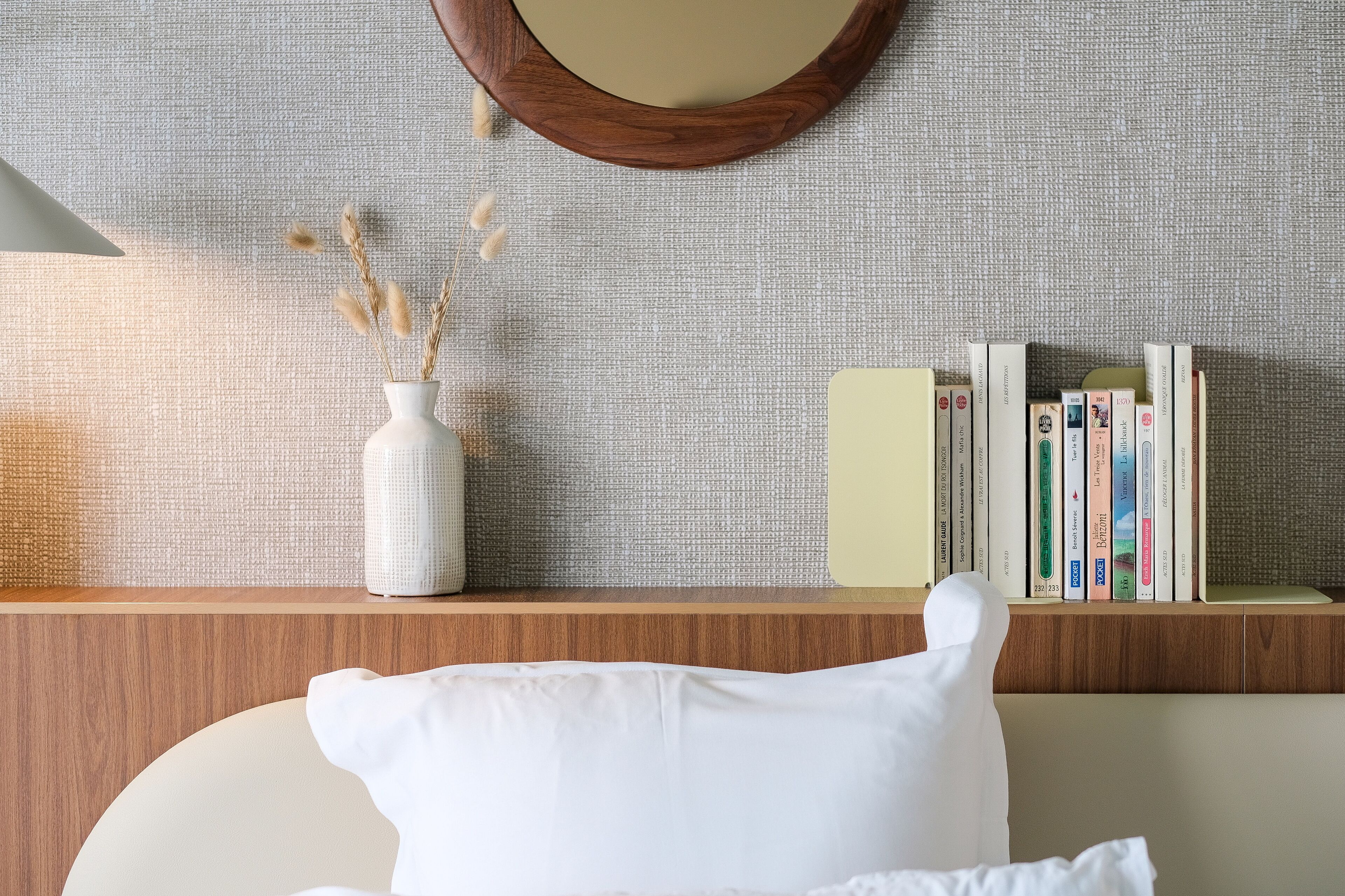 A neatly arranged bookshelf with books and a white vase with dried plants on a textured wall.