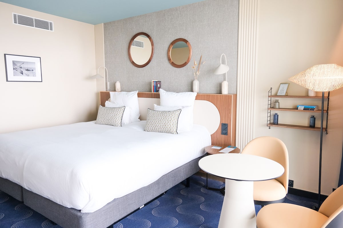 Una habitación de hotel moderna con una cama grande, una mesa redonda y una silla. Hay dos espejos en la pared, una estantería y una lámpara.
