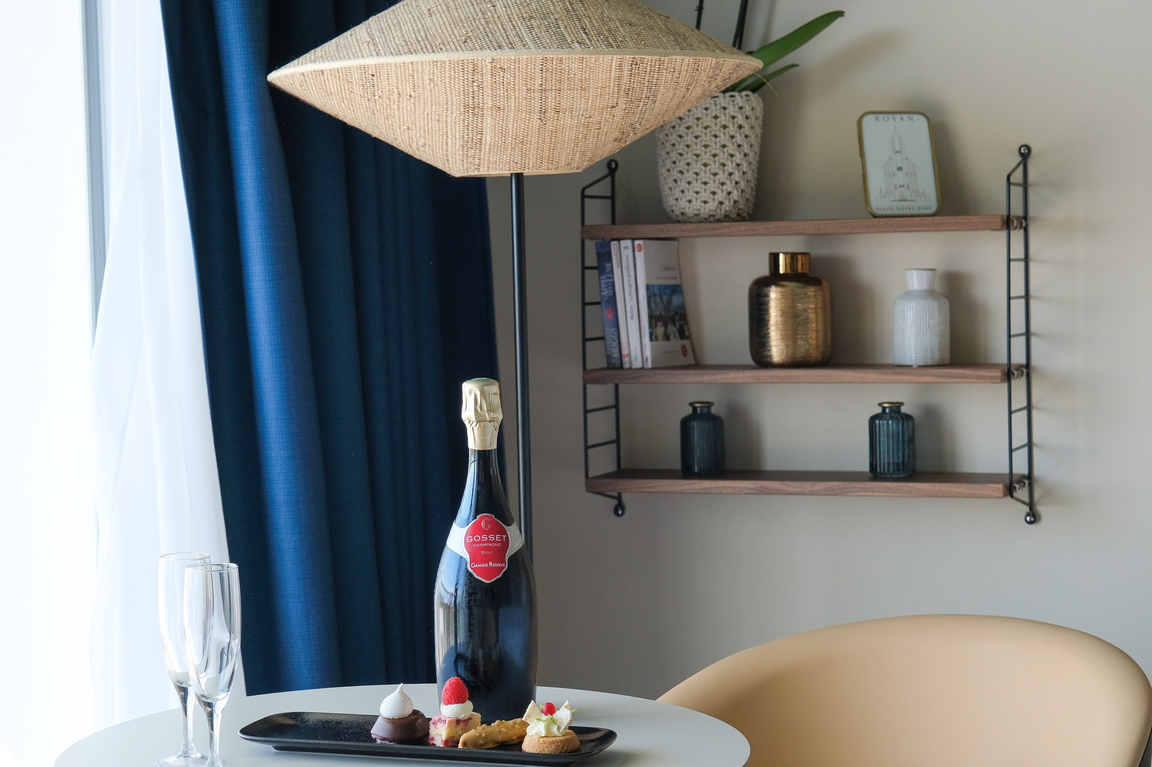 Une pièce avec une table ronde, une bouteille de champagne, deux verres et un plateau de desserts. On y trouve également une lampe suspendue, une étagère avec des livres et des objets décoratifs, et une chaise.