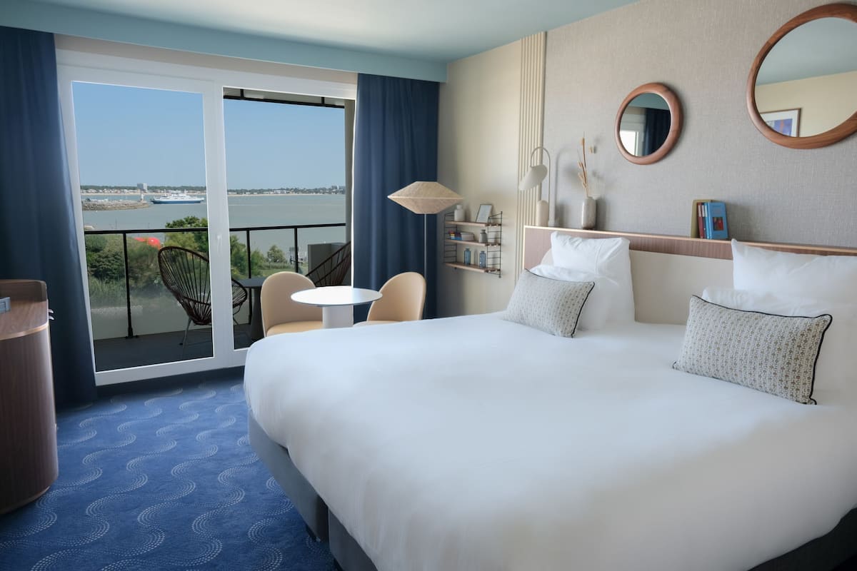 Habitación de hotel con una cama grande, una mesita, dos sillas, un balcón con vistas a un río y al paisaje urbano, y una estantería con objetos decorativos.