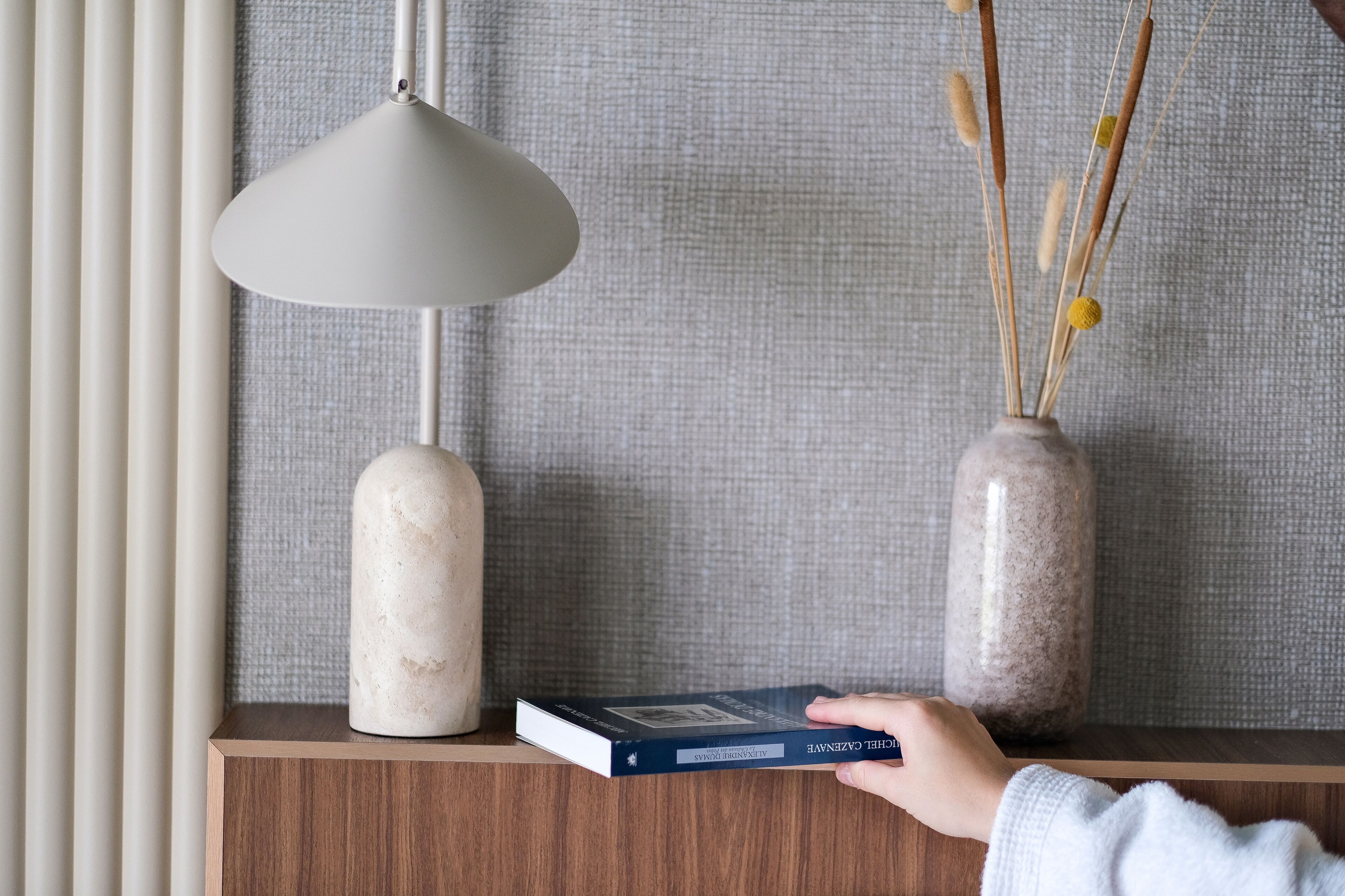 A book titled "Alexander Graham Bell" on a wooden shelf, a white vase with dried reeds, and a grey pendant lamp.