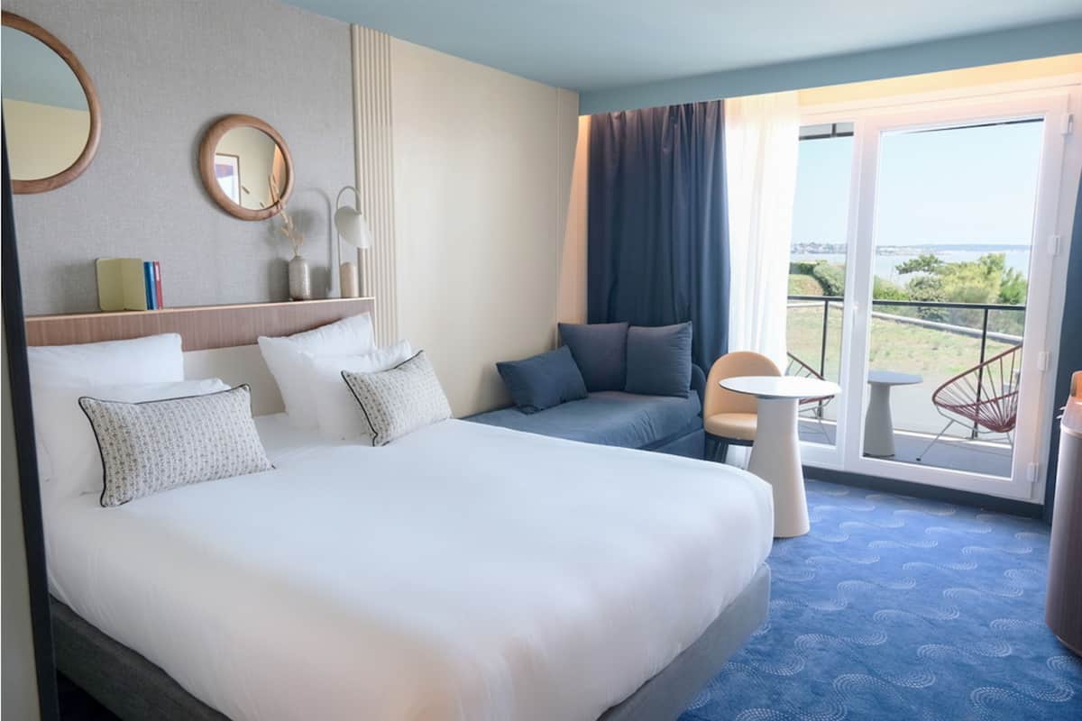 Una habitación de hotel con una cama grande, un sofá, una mesita y una silla. Tiene un balcón con vistas al mar.
