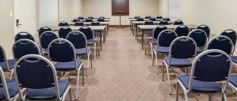 A conference room with rows of tables and chairs, a projector screen, and a carpeted floor.