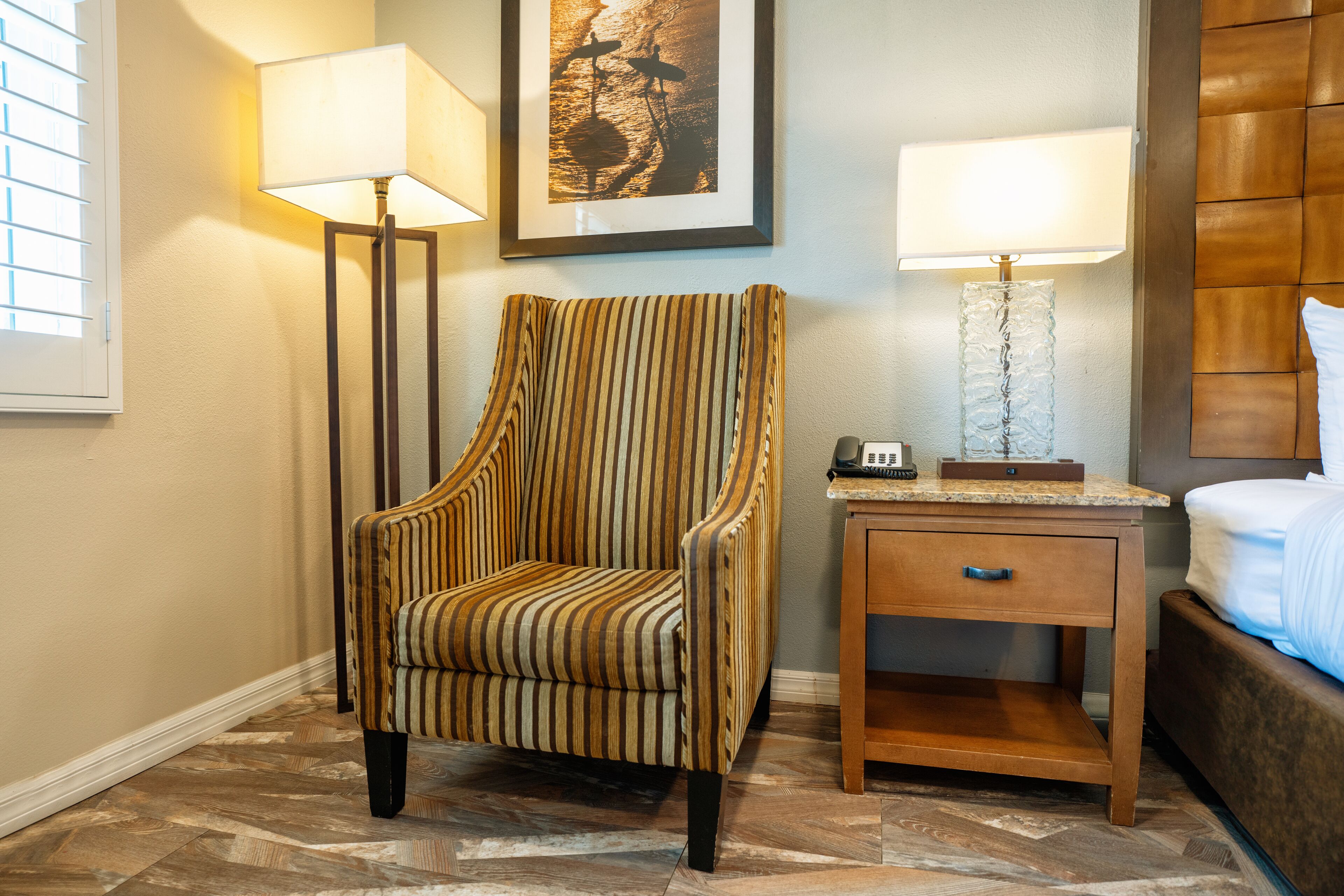 A hotel room with a striped armchair, a nightstand with a lamp, a framed picture on the wall, and a bed with a headboard.