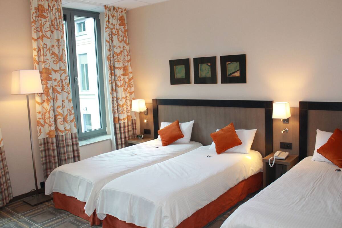 A hotel room with two beds, a window with curtains, wall-mounted lamps, and framed pictures.