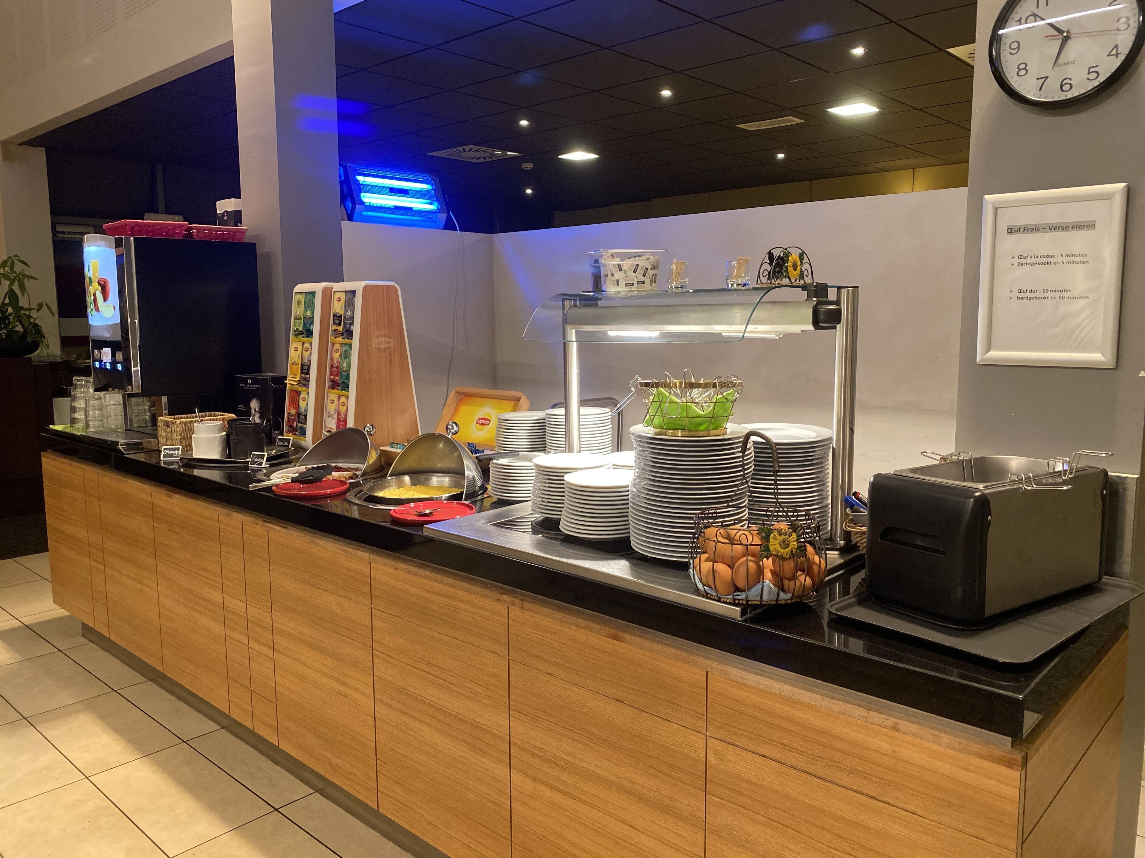 Un mostrador de buffet con diferentes alimentos, platos apilados y un reloj en la pared.