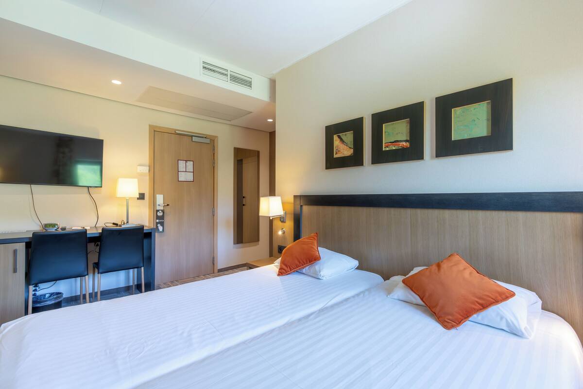 A hotel room with a bed, two chairs, a desk, a TV, and artwork on the wall.