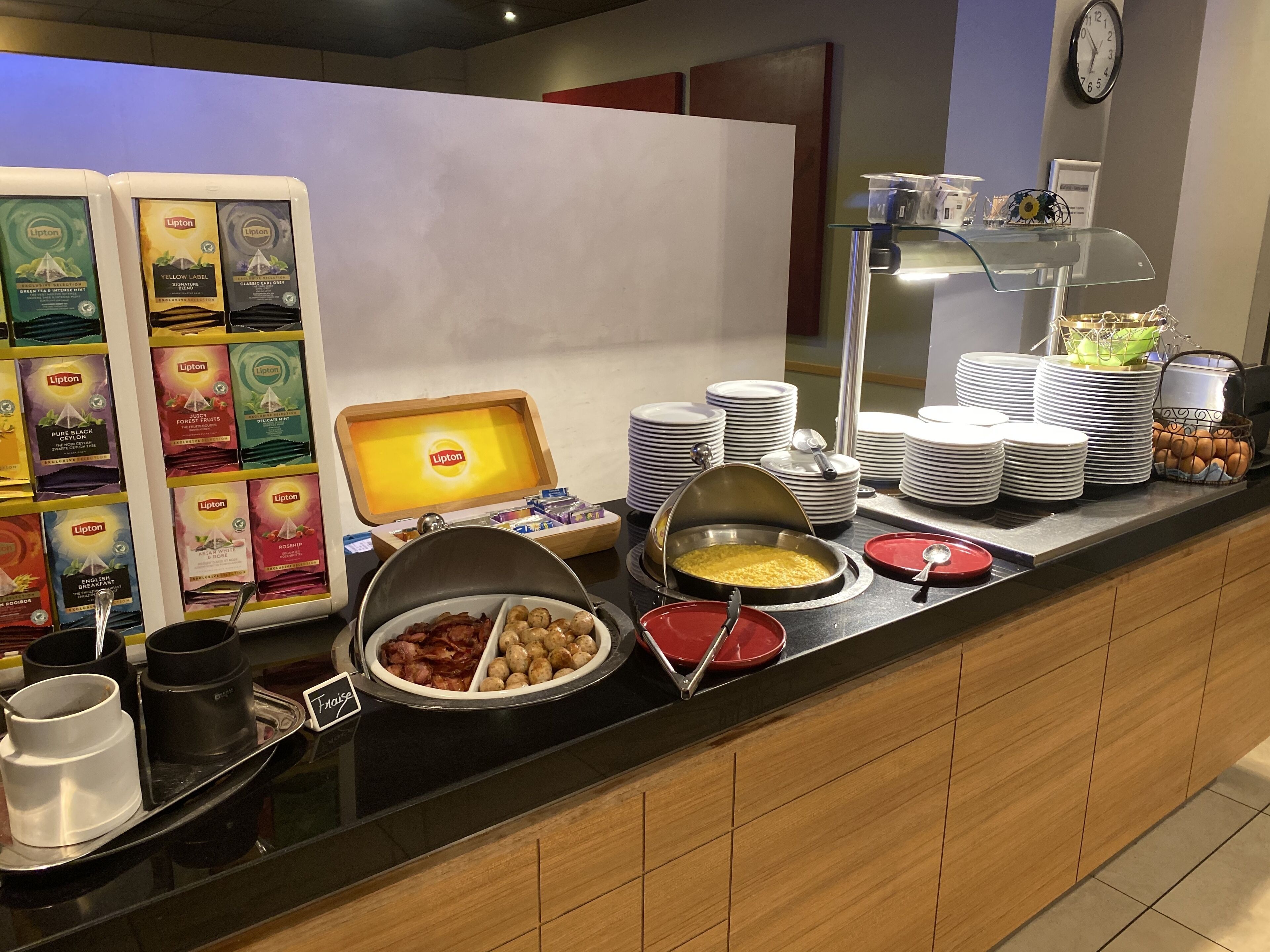 A buffet counter with various food items, including a bowl of bacon, a bowl of eggs, and a container of cereal. There are also stacks of plates and bowls on the counter.