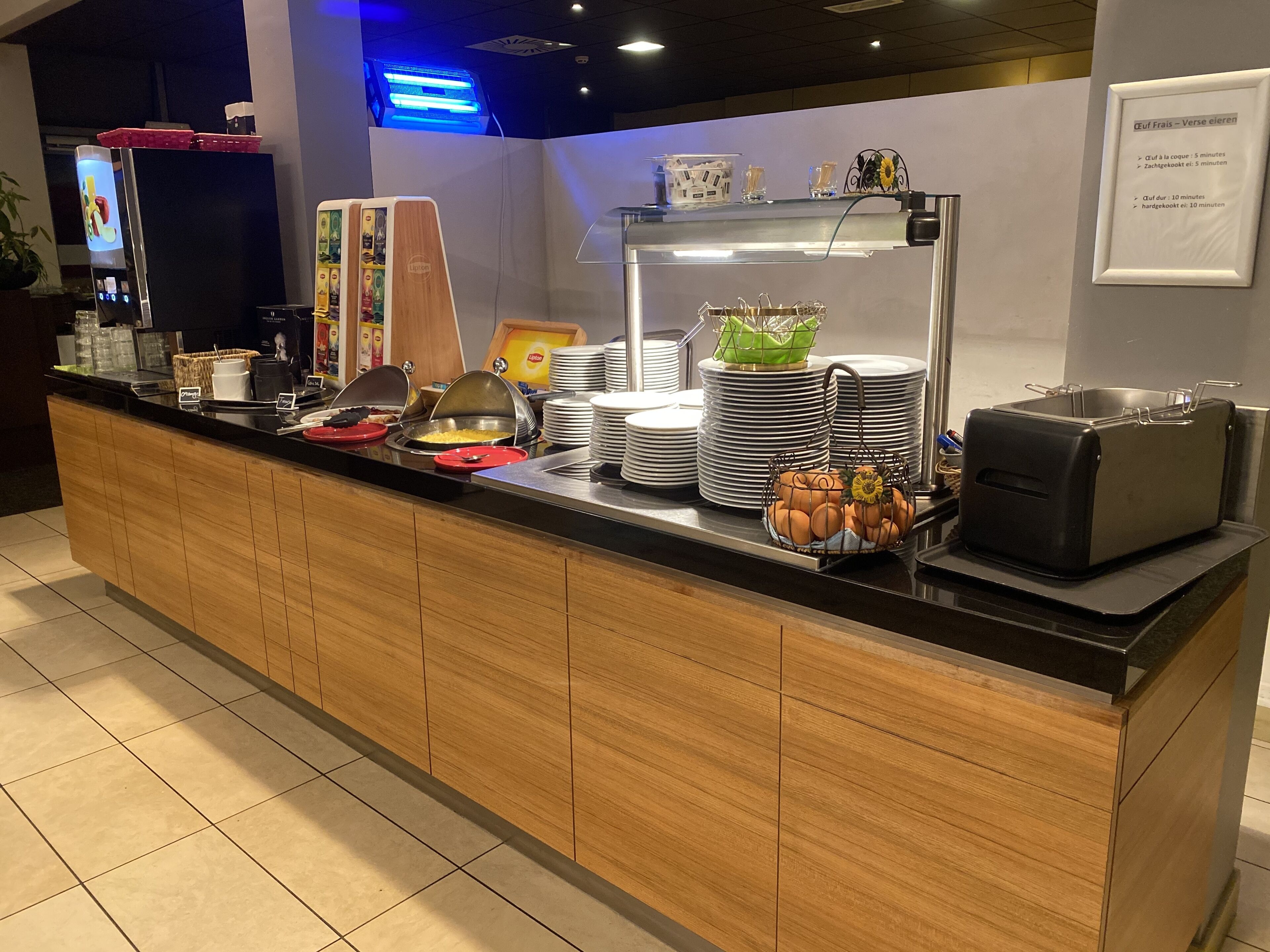 Een buffet met diverse gerechten, bestek en servies, een koffieapparaat, en een bord met openingstijden.