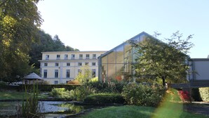 A building with a glass facade, a pond, and surrounding greenery. - Castel de Pont-à-Lesse Hotel (Dinant)